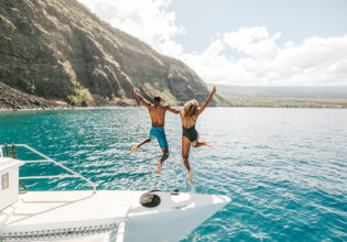 Two people jumping off a yacht with the beautiful coastline of Hawai‘i in the background.
