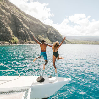 Two people jumping off a yacht with the beautiful coastline of Hawai‘i in the background.