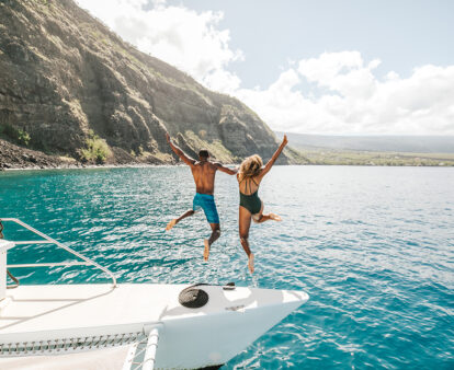 Two people jumping off a yacht with the beautiful coastline of Hawai‘i in the background.