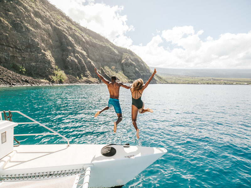 Two people jumping off a yacht with the beautiful coastline of Hawai‘i in the background.